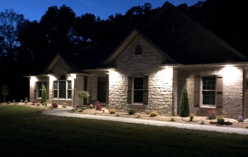Stone home lit by landscape lighting at dusk with flowering beds along the walk