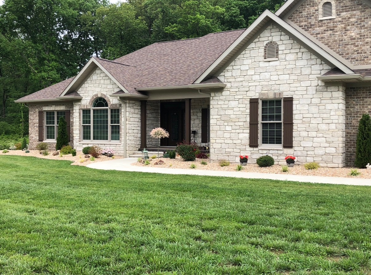 Stone home with a curved concrete walkway, layered beds and a manicured lawn